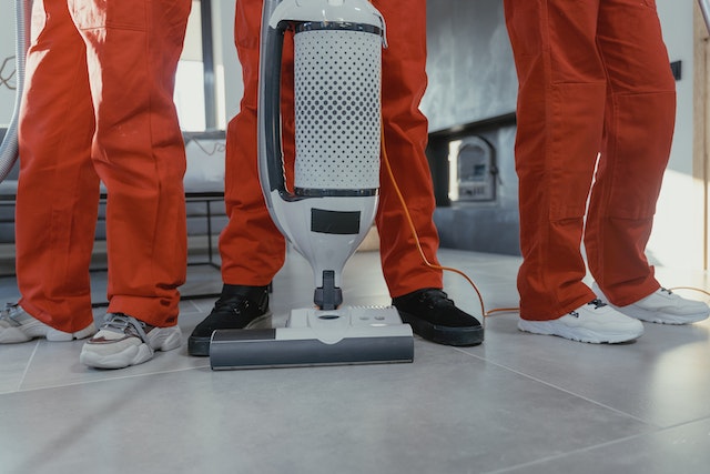 The legs of three people wearing orange pants behind a vacuum